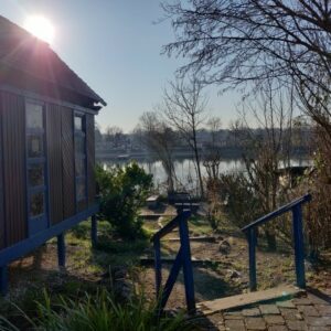 Man sieht die Location für den Outdoor-Workshop in Mannheim vom Gestaltcamp. Die Sonne scheint auf den Stollenwörthweiher in Mannheim, man sieht ein Lagerfeuer und den Pavillon aus Holz als Unterschlupf bei Regen