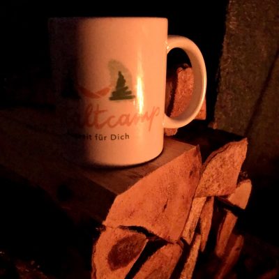 Gestaltcamp Tasse am Lagerfeuer bei einer gelungenen Veranstaltung: Winter Wunder Weg von Gestalttherapeutin Jennifer Schumacher-Hetzel, Outdoor Workshop am 18.12.22 in Mannheim zu sehen ist eine Tasse mit dem Logo des Gestaltcamps in Mannheim auf Holzscheiten, die angeleuchtet wird von dem Licht eines romantischen Lagerfeuers. Die tolle Tasse und die schöne Erinnerung an den Abend kann man im Gestaltcamp-Shop kaufen. Workshops Outdoor in Mannheim z.B. zur Wut oder zur Erholung "Akku Aufladen" finden am Stollenwörthweiher statt.