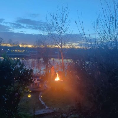 Teilnehmerin eines Workshops am Lagerfeuer vor dem Stollenwörthweiher in Mannheim im stadtökologischem Zentrum Auf dem Foto sieht man eine Frau mit einem Stockbrot, die am Lagerfeuer steht. Im Hintergrund sieht man den Veranstaltungsort, den Stollenwörthweiher in Mannheim und dahinter die gelb leuchtende untergehende Sonne. Eine schöne Veranstaltung mit viel Atmosphäre in der freien Natur. Workshops Outdoor in Mannheim auf Basis der Gestalttherapie,