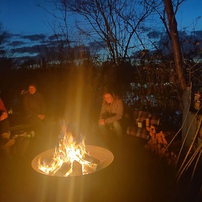 Drei Frauen sitzen gemütlich in romantischer Lagerfeuer-Atmosphäre gesellig beisammen und schauen in die knisternden Flammen. Im Hintergrund ist ein sternenklarer Himmel in Mannheim am See zu sehen. Werbung für den Workshop Kleine Visionssuche am Lagerfeuer mit Julie Strobach am 28.05.22 in Mannheim am Stollwörthweiher - Outdoor vom Gestaltcamp